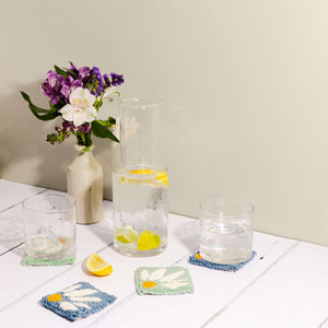 Punch needle coaster in blue and mint green, lemon water in a jug with glasses atop the coasters. A slice of lemon lies in the foreground.