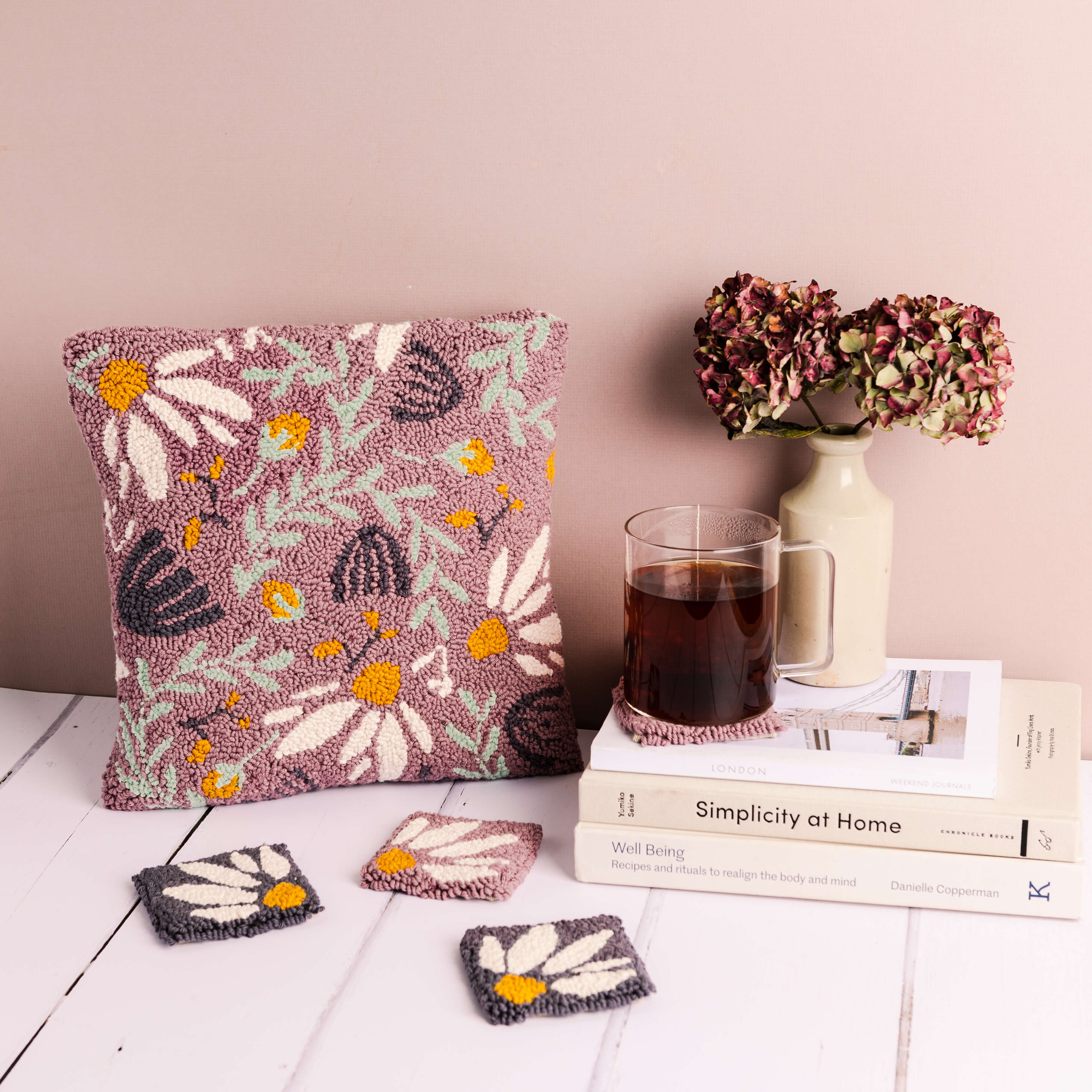Lilac floral punch needle cushion against a pink background with punch needle coasters in the foreground. There are books with a vase and glass cup of tea to the right.