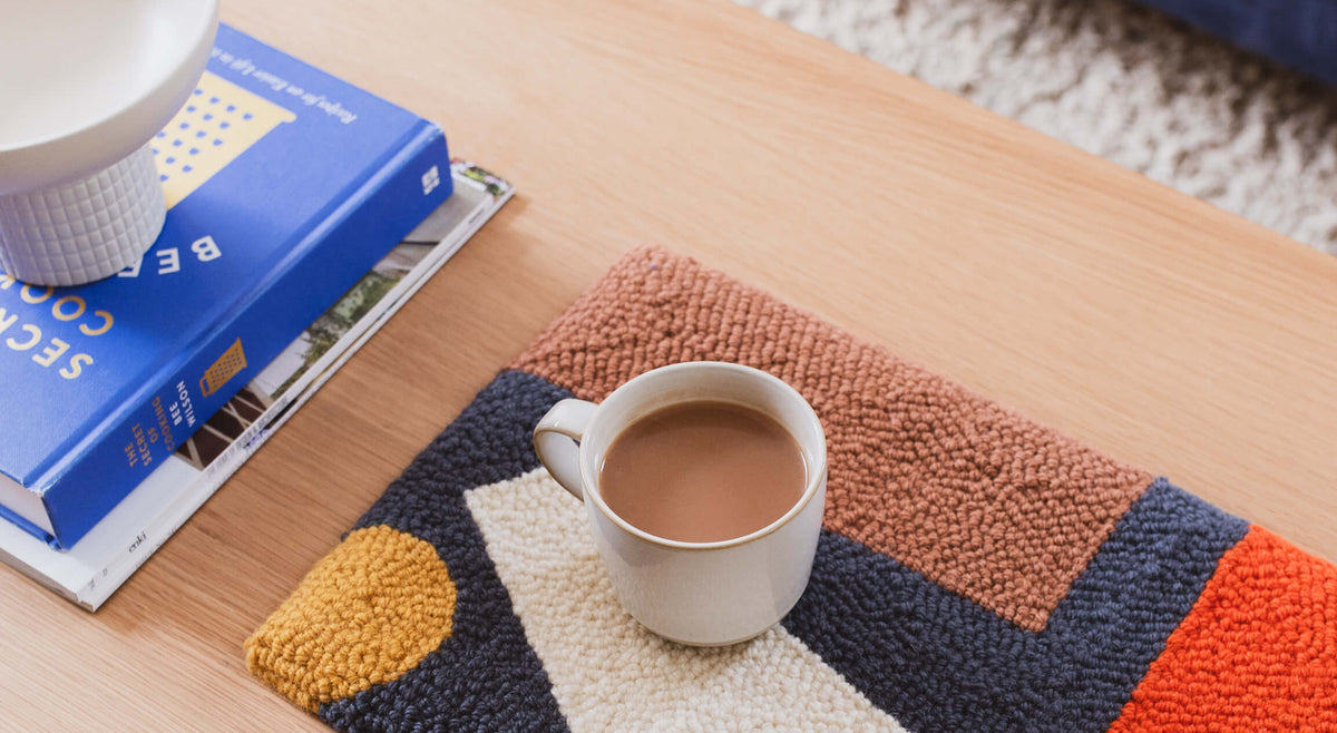 A rectangular abstract-patterned rug in navy, cream, rust, orange, and mustard sits on a light wood coffee table. A white mug filled with coffee rests on the rug, with books and a blue chair visible in a styled indoor setting.