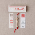 Oxford Punch Needle kit laid out on a beige linen background, including the wooden punch needle tool with red thread, a white branded box, a stitch gauge card, and a quick guide card.