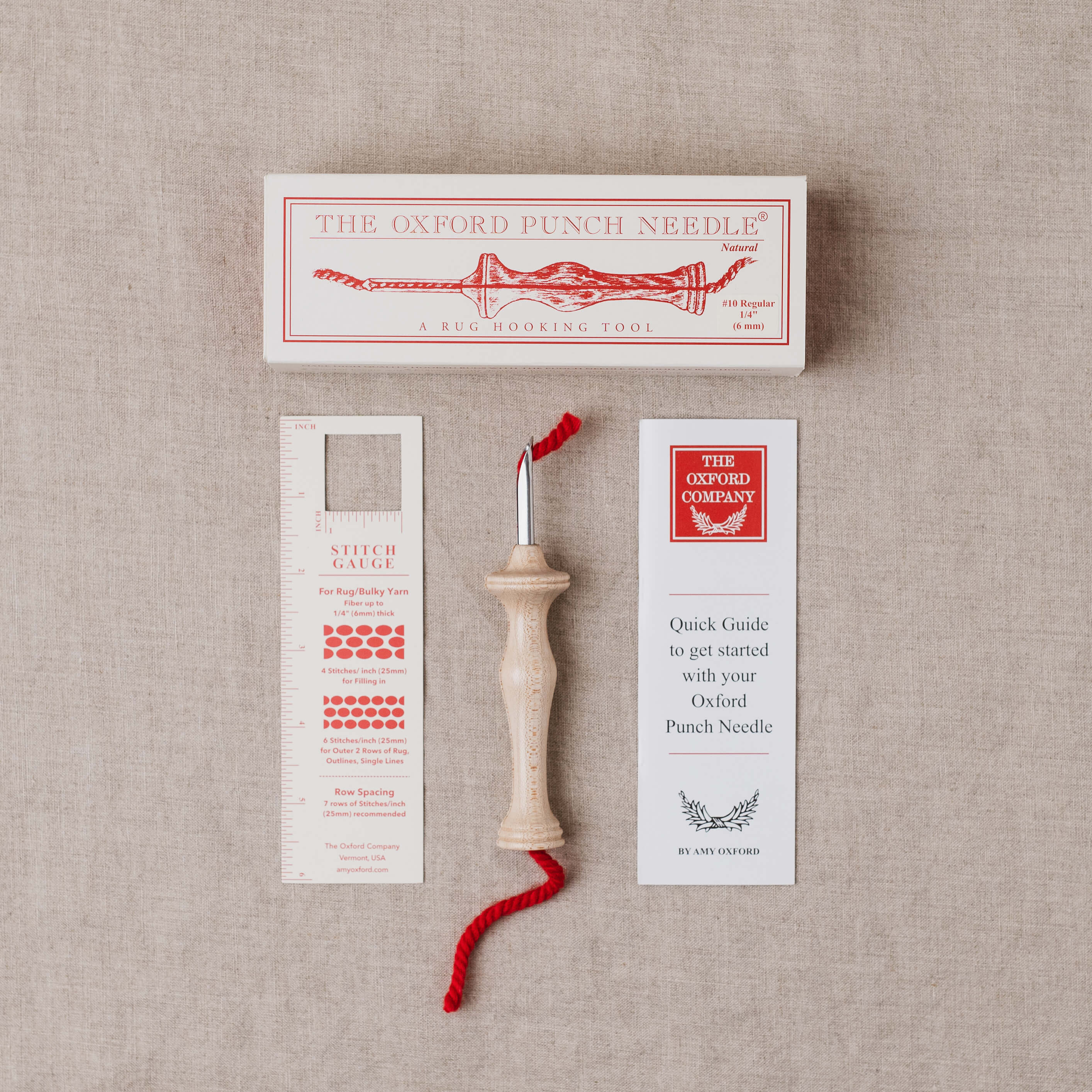 Oxford Punch Needle kit laid out on a beige linen background, including the wooden punch needle tool with red thread, a white branded box, a stitch gauge card, and a quick guide card.