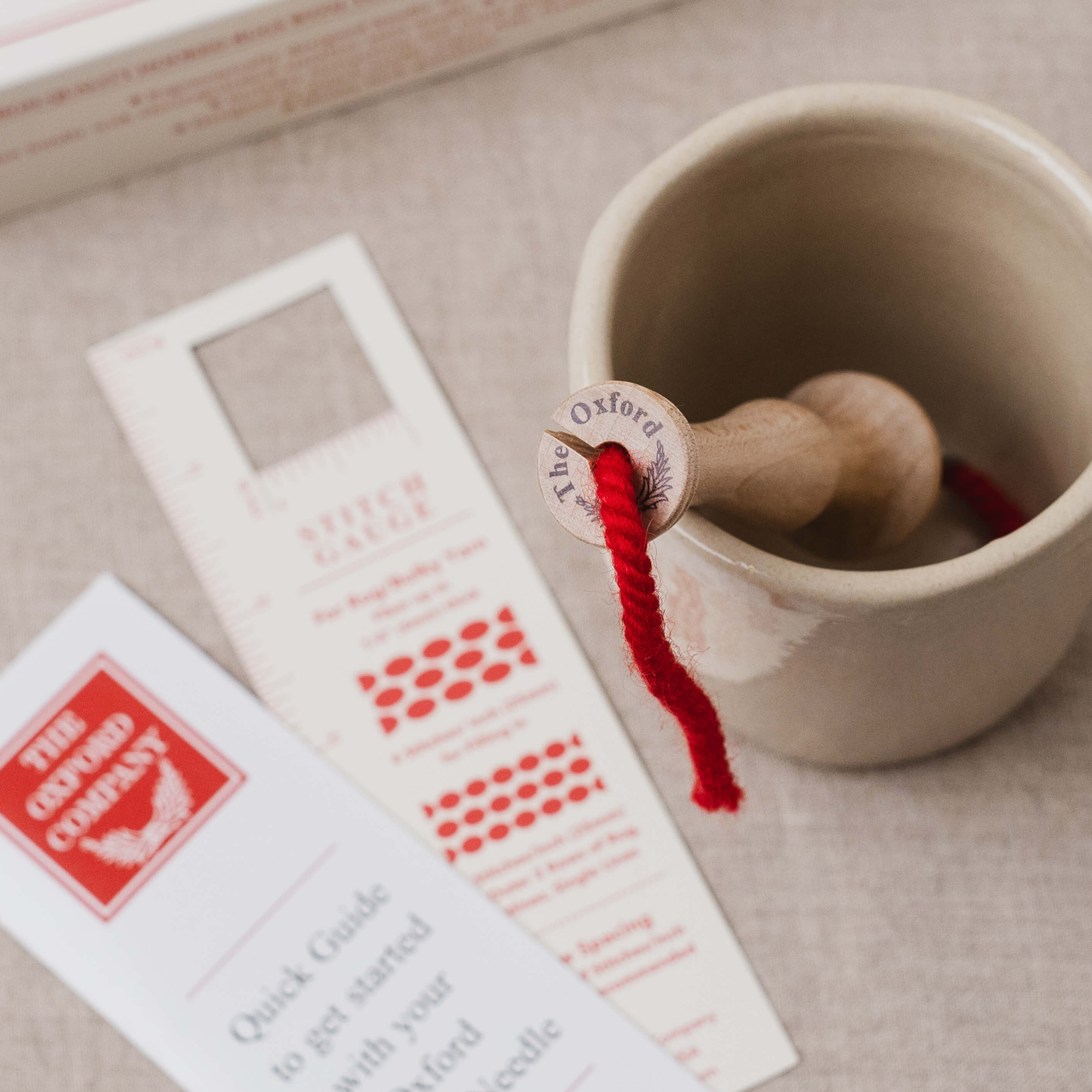 oxford punch needle in ceramic cup with red yarn extending, stitch gauge and quick start guide lay beside it