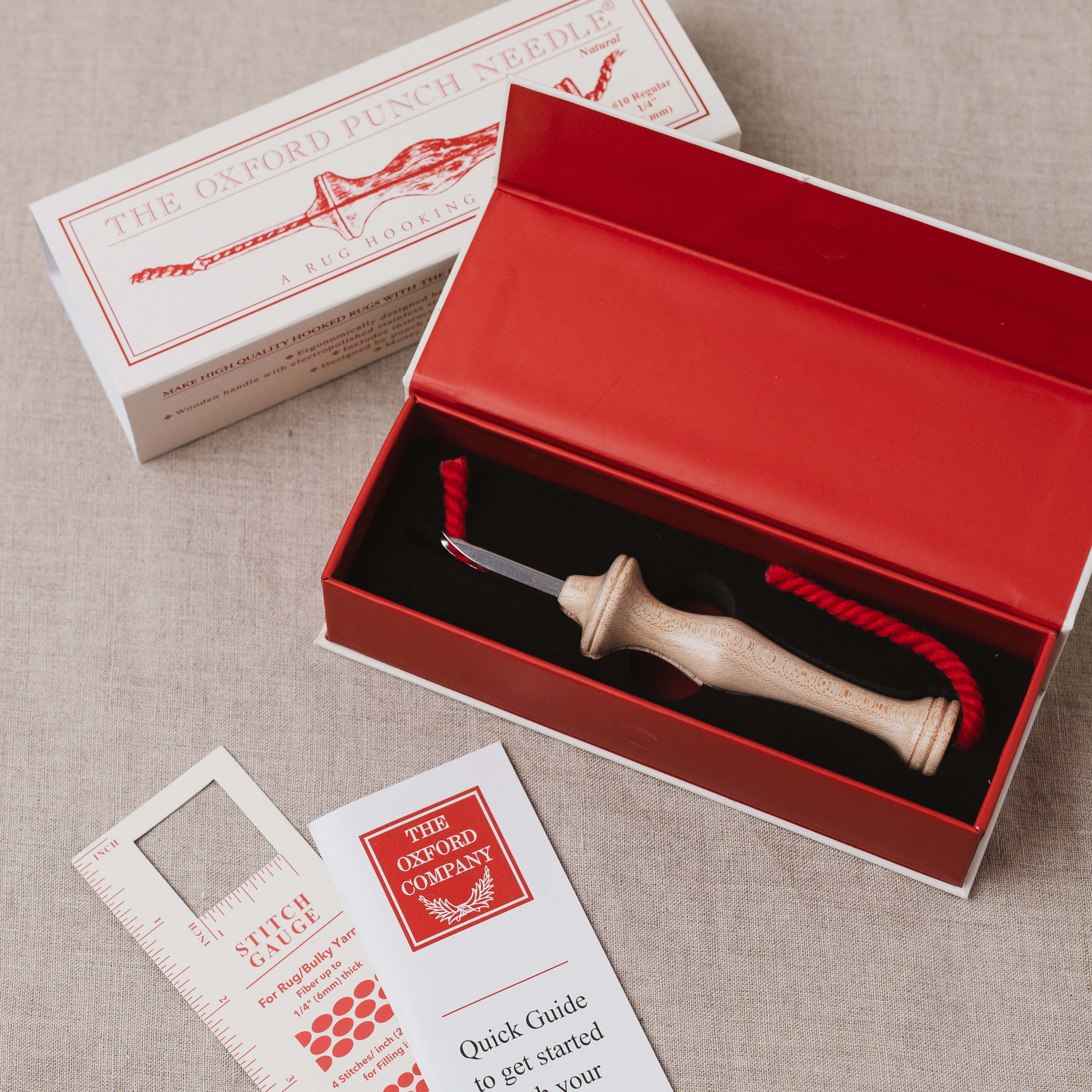 The Oxford Punch Needle kit shown with its red presentation box, white branded box lid, instruction booklet, and a wooden punch needle tool with red cord on a neutral fabric background.
