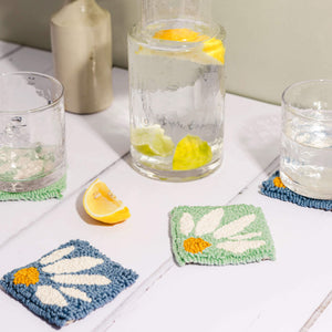 Punch needle coaster in blue and mint green, lemon water in a jug with glasses atop the coasters. A slice of lemon lies in the foreground.