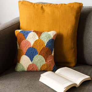 Scallop punch needle cushion in autumnal 70's tones propped on a sofa with a burnt orange cushion behind and book open