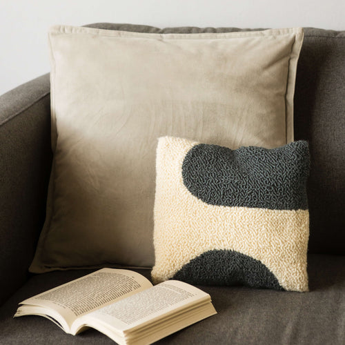 Grey and white shapes punch needle cushion propped on sofa with larger grey cushion and open book